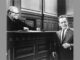 A black and white photo with an judge with glasses seated at the bench sternly looking at a man in a suit and tie that seems to making a case.