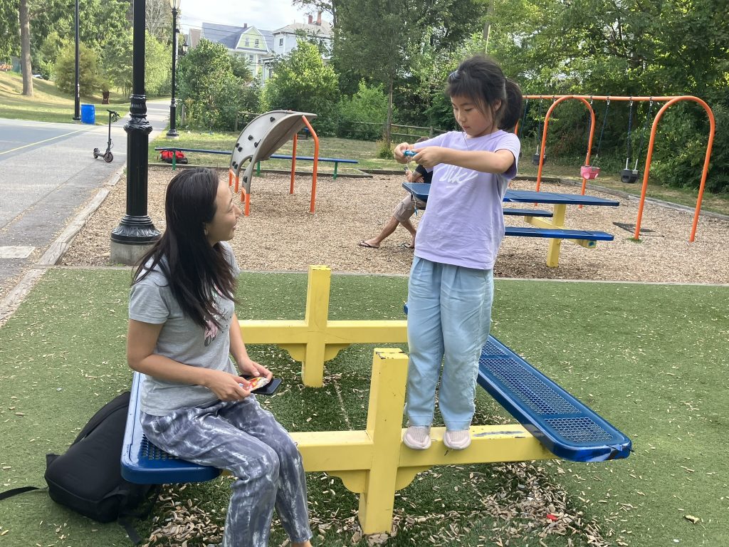 A young mom with long black hair sitting on a colored picnic bench smiling at her pre-teen daughter who stands on the other side playing with a string toy 