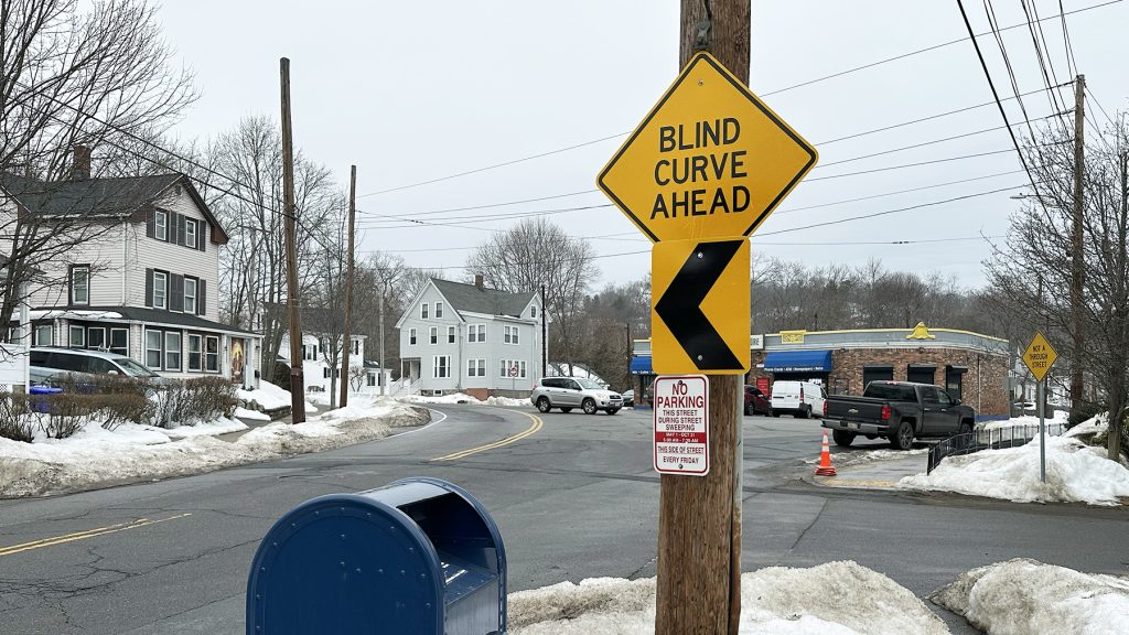 Intersection showing "blind curve ahead" sign, streets, and store, as described in caption.
