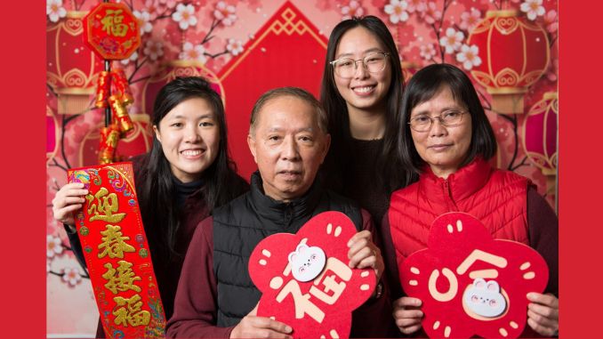 An Asian family of four with two young adult children - looking straight at the camera holding red hearts decorated with Chinese symbols and surrounded by red decorations