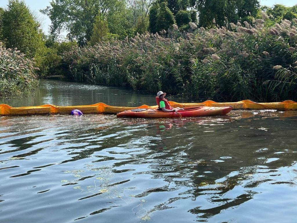 A person in a kayak wearing a hat on a river next to an orange floating ridge which is the trash trap.